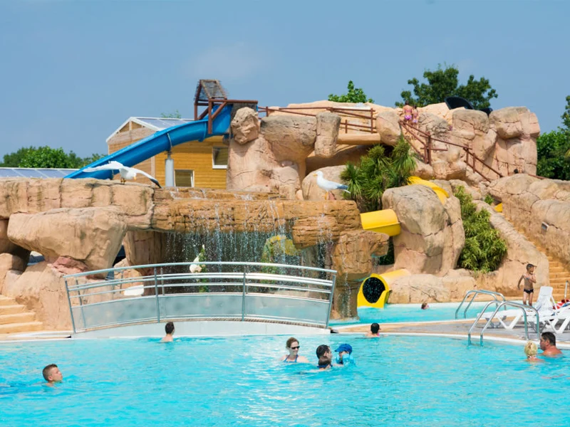 Outdoor pool with people swimming; waterfall feature flows from faux rocky landscape with blue and yellow slides at Le Front de Mer