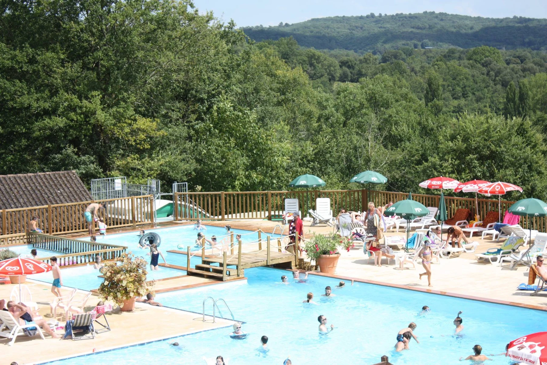 Swimmers enjoying pools, people relaxing on loungers under umbrellas, surrounded by lush trees and distant hills at Les Grottes De Roffy