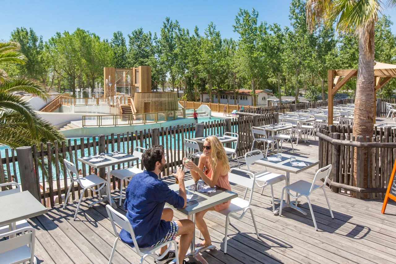 Couple dining outdoors with drinks, surrounded by trees and a pool at Blue Bayou