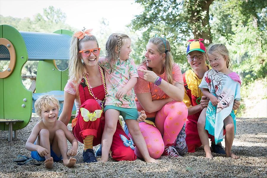 Adults and children dressed colorfully play and smile together in a playground surrounded by trees at Slow Village Provence Occitane 