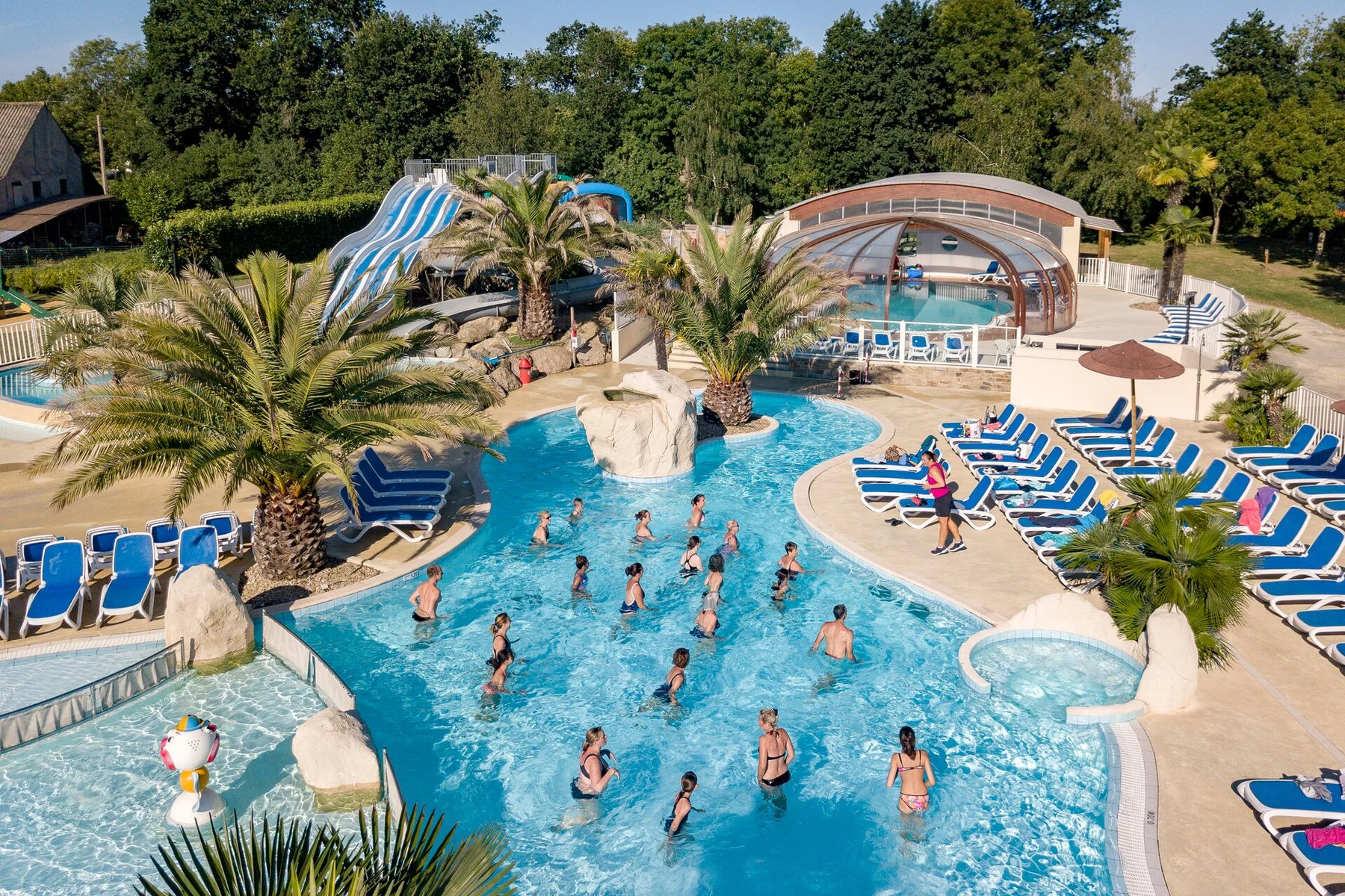 People exercising in a large outdoor pool surrounded by palm trees and lounge chairs at Les 2 Fontaines