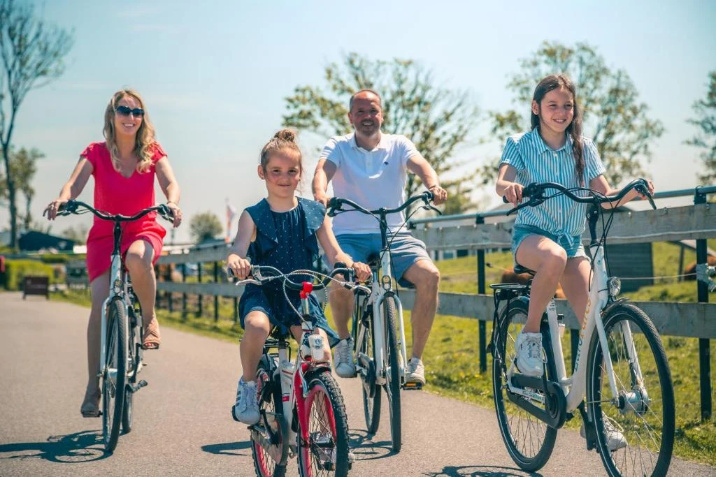Family biking together on a sunny path, surrounded by green grass, trees, and a wooden fence at Europarcs de Rijp