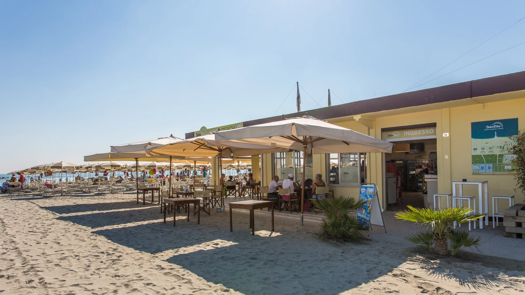 People sitting under large umbrellas at an open-air beach cafe, with sandy ground and nearby ocean waves at Pini Beach Village 
