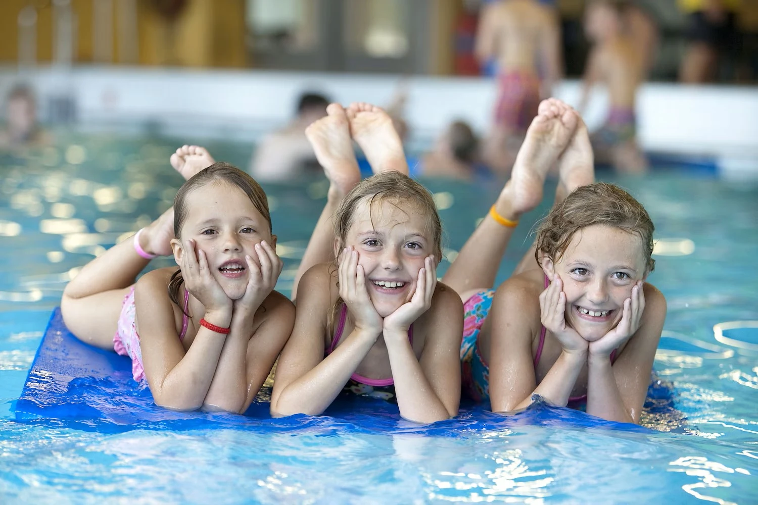 Three children lying on a blue float in a pool, smiling with hands under chins; other swimmers in background at Bospark De Schaapskooi