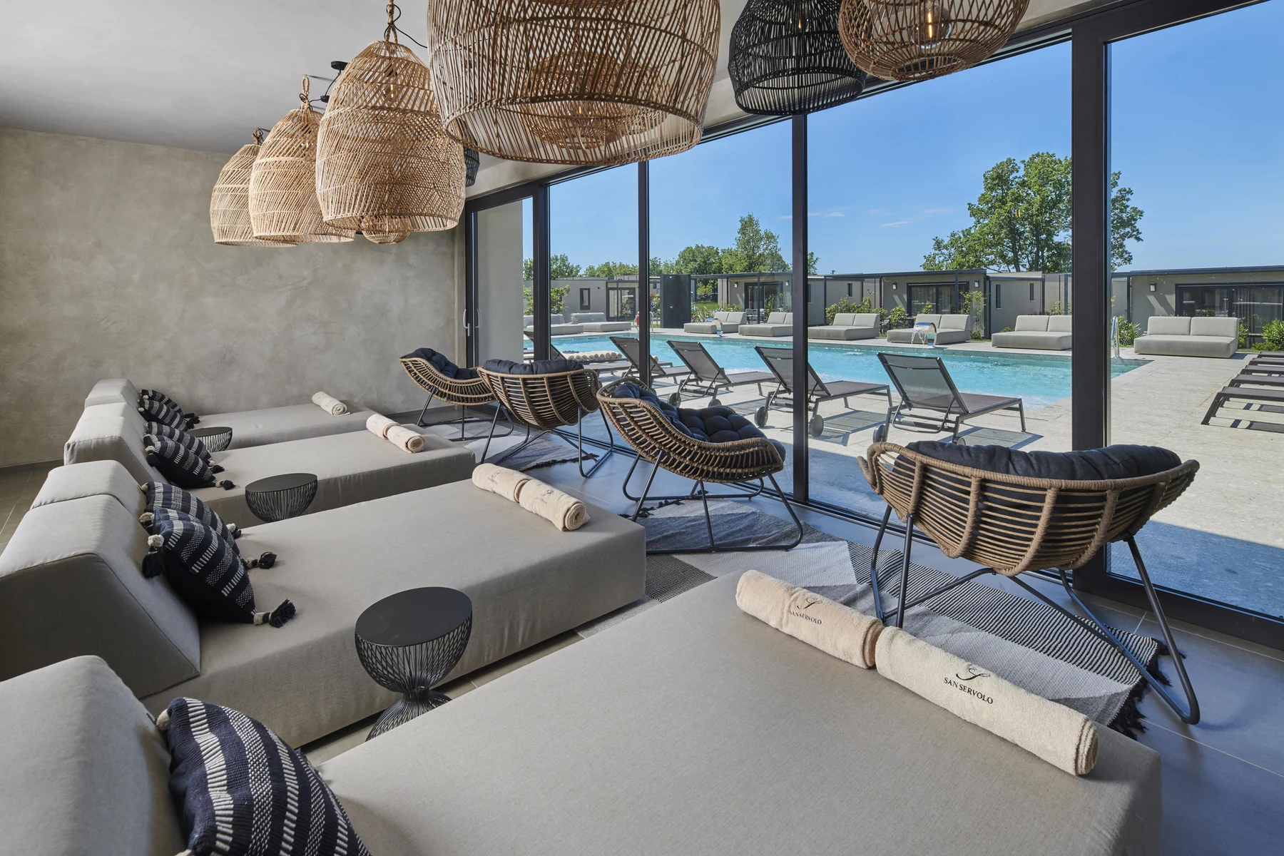 Loungers and wicker chairs arranged by a large window overlook a swimming pool in a resort setting at San Servolo Resort