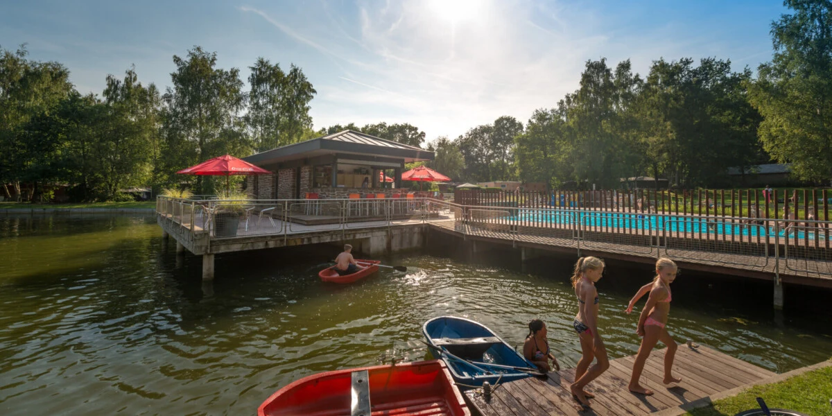 Children swimming and kayaking in a lake near a wooden deck with a cafe adorned with red umbrellas, surrounded by trees at Jocomo Park