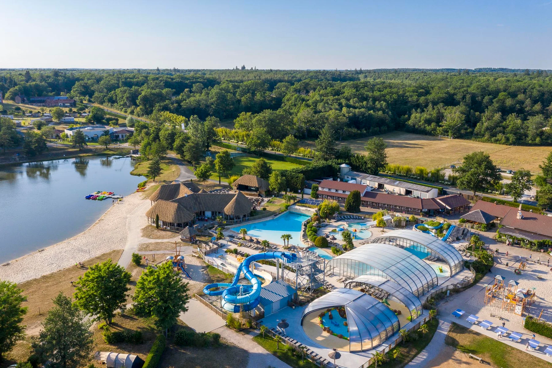 Waterpark surrounded by cabins, sandy beach, trees, a small lake, and residential buildings in a forested area at Les Alicourts Resort