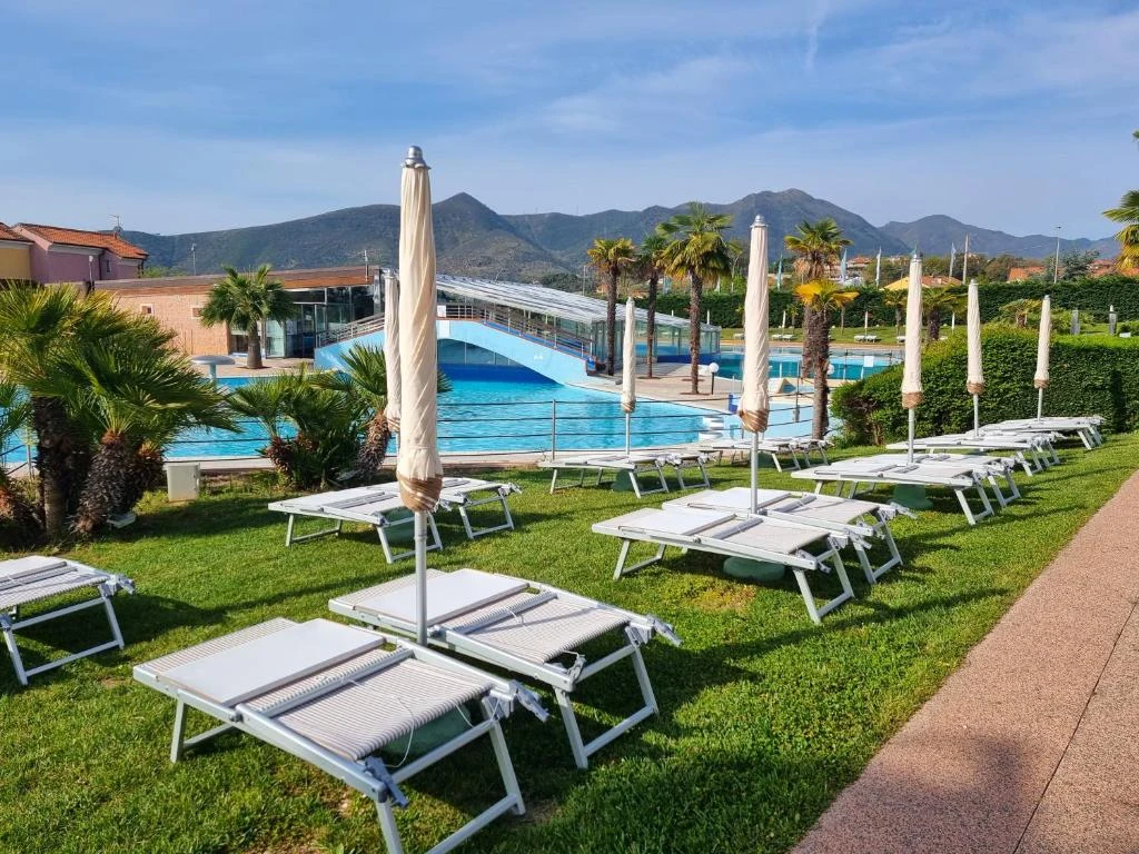 Lounge chairs with closed umbrellas are arranged on grass near a swimming pool, with mountains in the background at Loano 2 Village