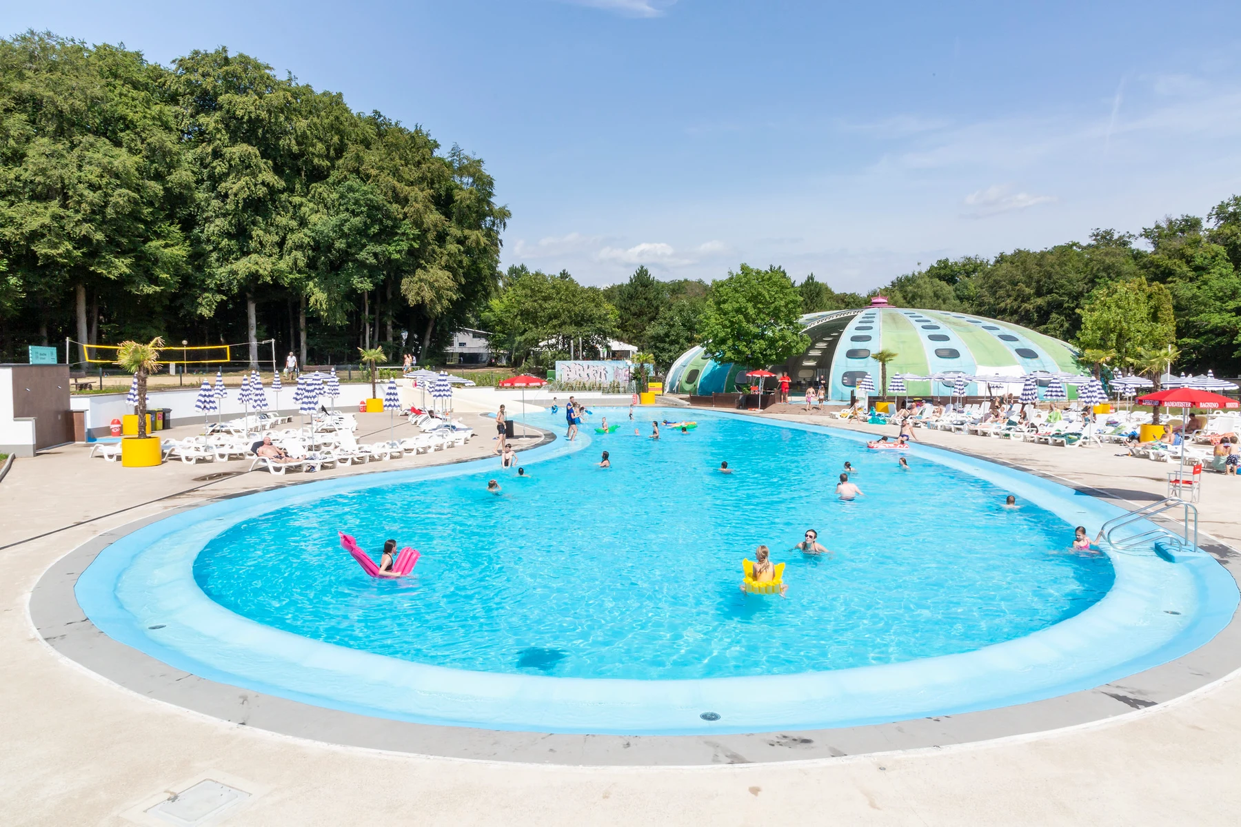 Swimming pool with people playing and relaxing, surrounded by loungers and umbrellas, near green dome structure in sunny forested area at hu Birkelt Village