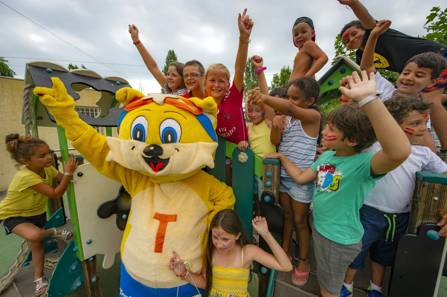 Character mascot posing with children on playground equipment, while all are smiling and raising their hands at Les Salins de la Gardiole 