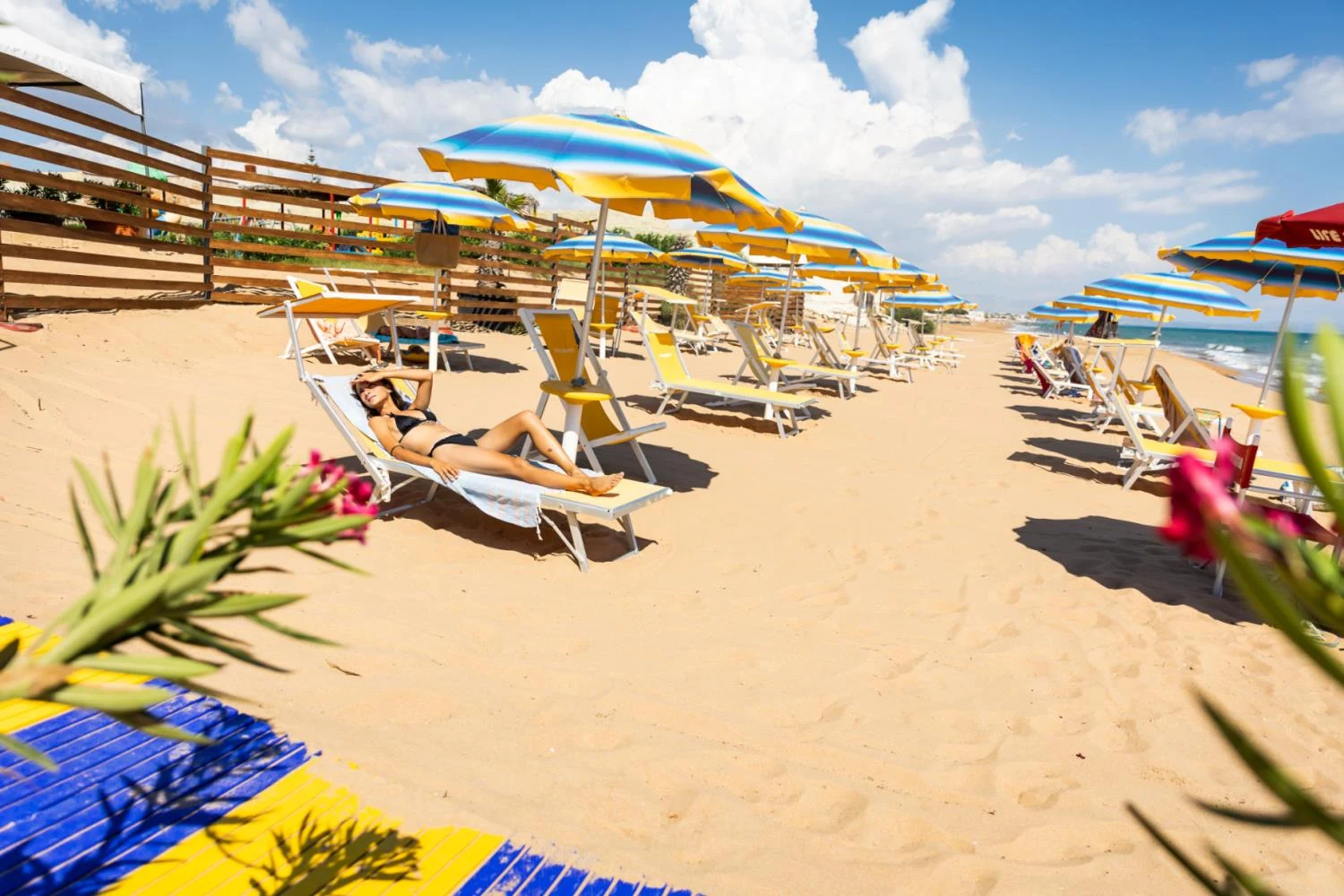 Woman sunbathing on a lounge chair under an umbrella on a sandy beach with rows of similar setups at Triscina Mare