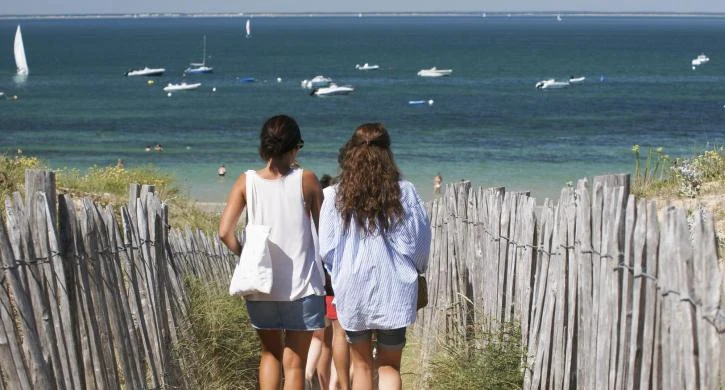 People walking down a sandy path between wooden fences towards a beach with boats on water at Seasonova Ile de Ré