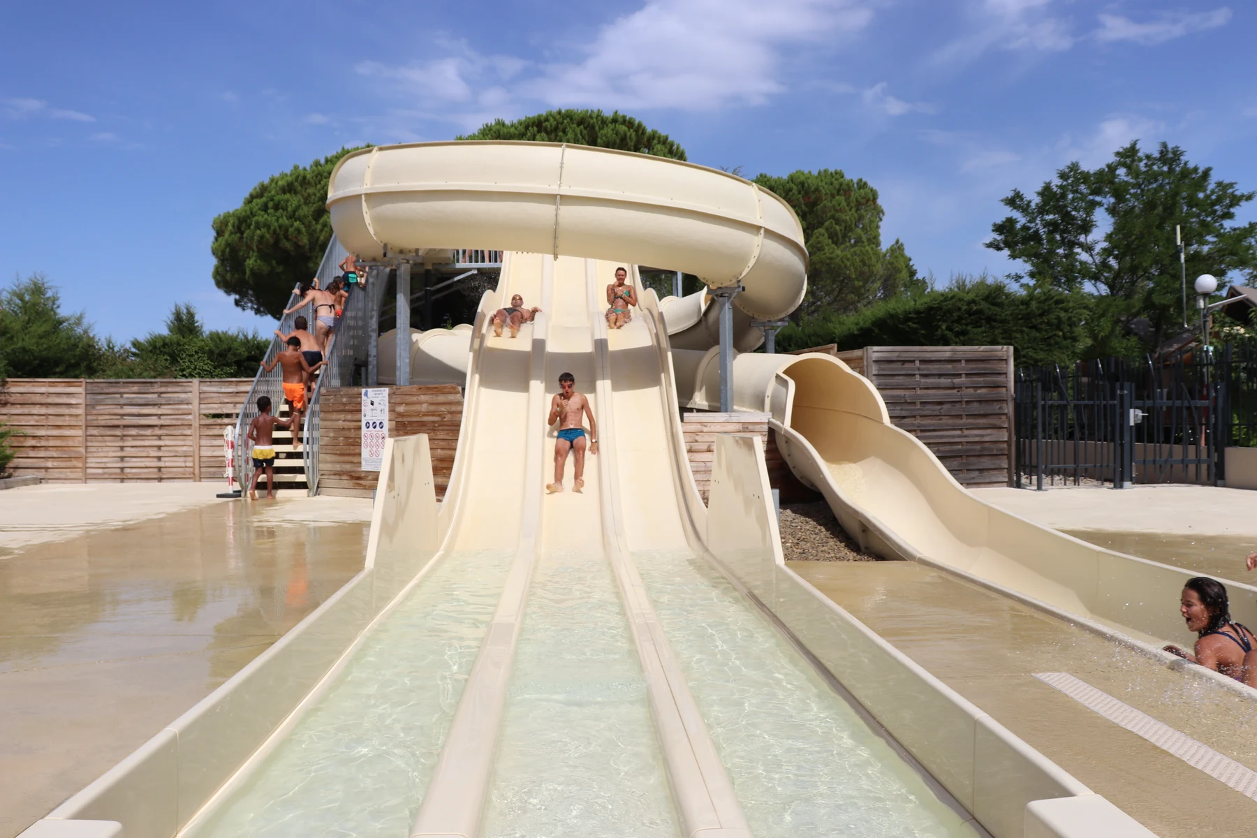 Water slides with people sliding down, surrounded by a wooden fence and trees, under a clear blue sky at International Aups
