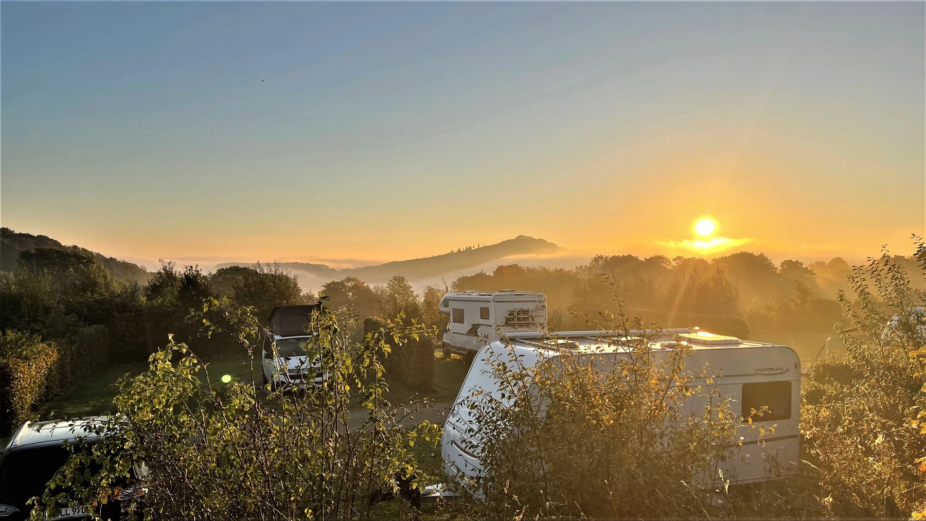 Caravans parked in a forested area during a scenic sunrise with misty hills in the background at Brilon