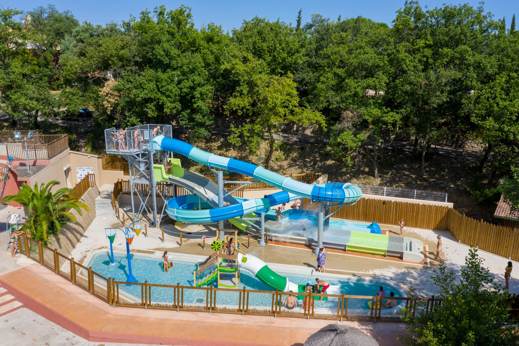 Waterslide spirals down into a pool with children playing and adults supervising, surrounded by trees and wooden fences at Le Bois Fleuri