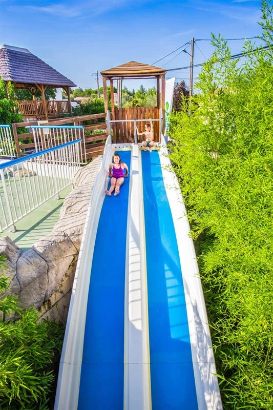 Two children sliding down side-by-side water slides surrounded by greenery and wooden structures at Le Petit Bois