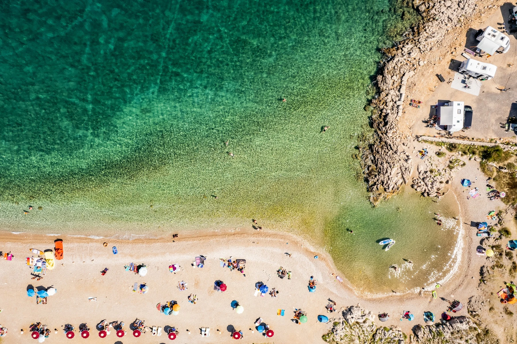 Beach crowded with sunbathers and umbrellas, people swimming in turquoise water, campers parked on rocky terrain; at Camp Tiha