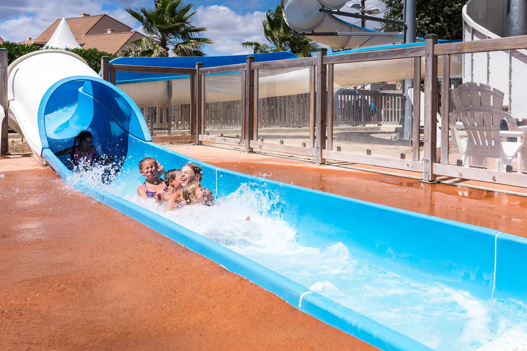Children slide down a blue water slide, splashing into a pool under a sunny sky at Marvilla Parks Dunes et Soleil