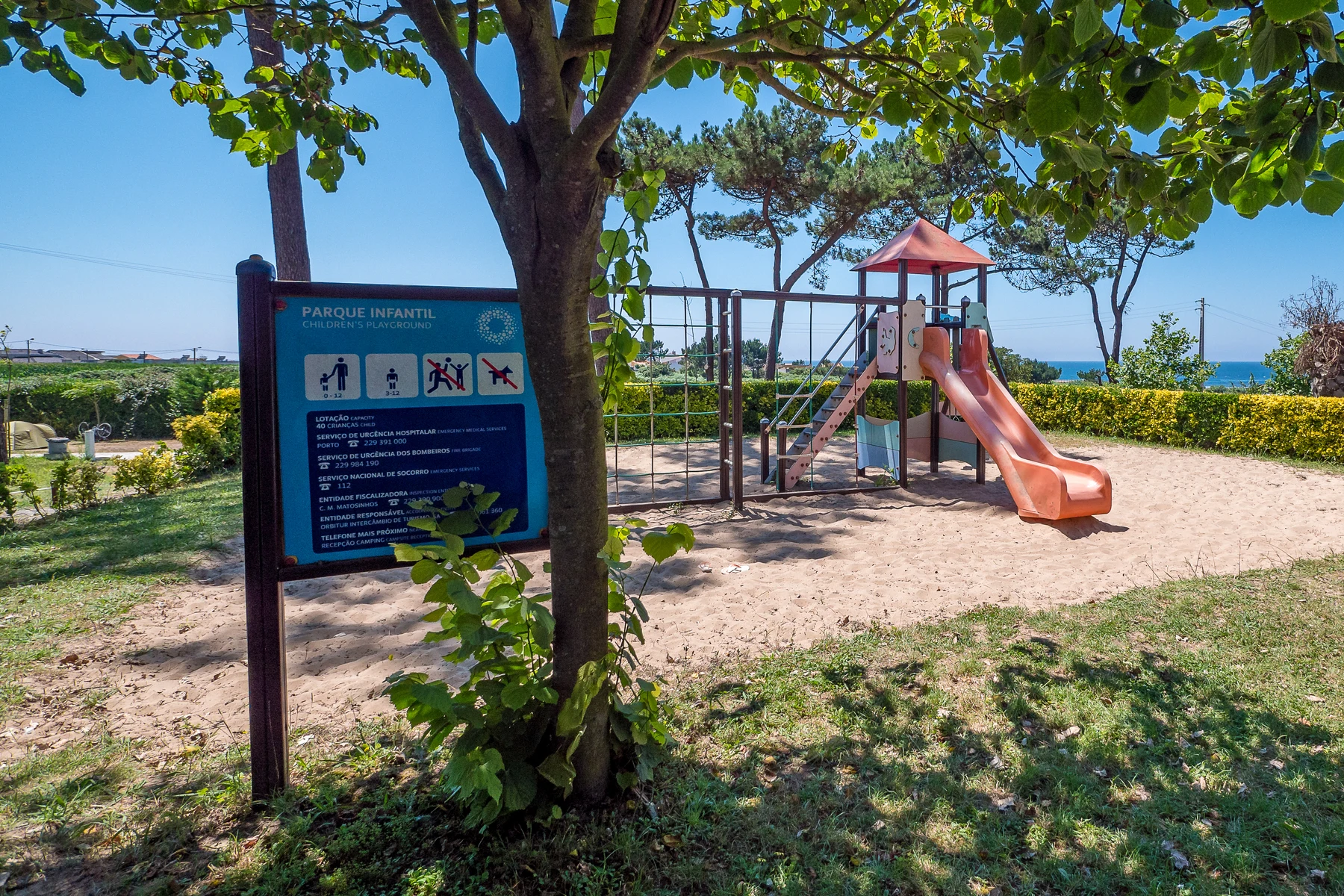 Playground equipment stands unused on sandy ground, surrounded by greenery and a signboard at Angeiras