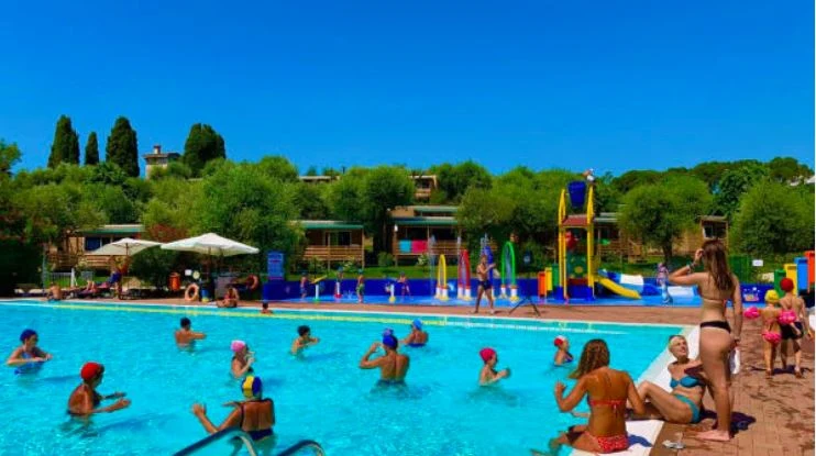 People swimming and relaxing in a large pool, with colorful slides in the background, surrounded by lush greenery at Camping Della Colombaia