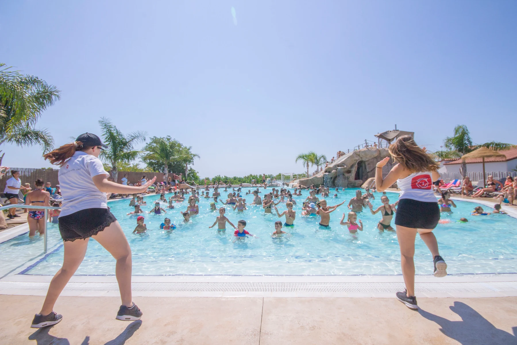People performing water aerobics led by two instructors near a large outdoor pool, surrounded by palm trees and a sunny sky at Alannia Els Prats