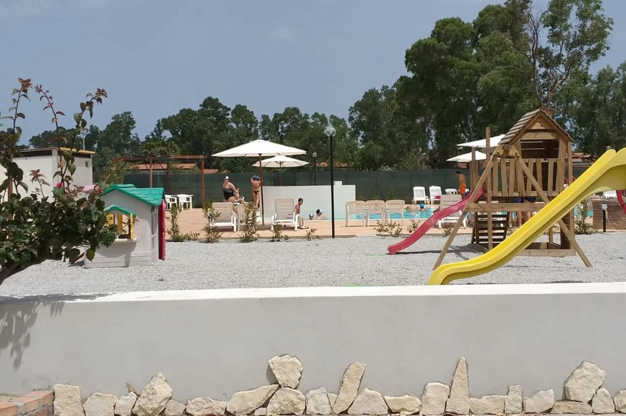 Wooden playground structure with slides next to a pool area with people relaxing under umbrellas; trees in background at Tindari Village
