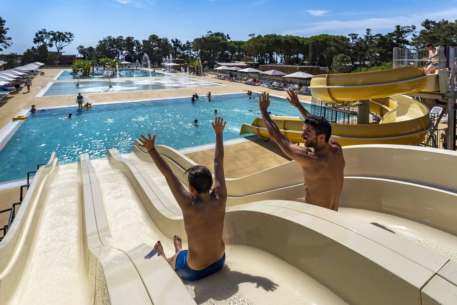 Two people slide down water slides into a large swimming pool area surrounded by lounge chairs and trees at Cap Sud