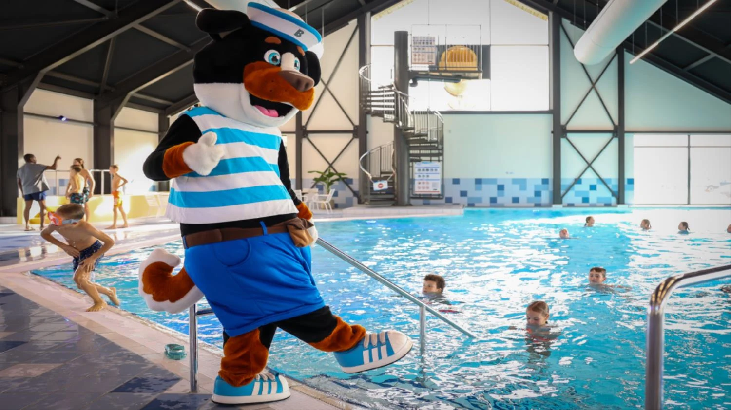 Mascot bear in sailor outfit standing by indoor swimming pool with kids swimming and playing nearby at Maasresidence Thorn