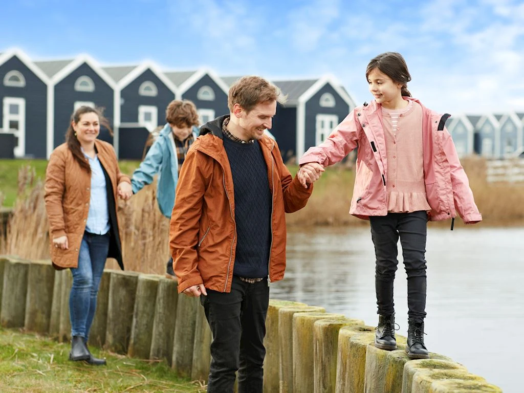 Child balancing on wooden beams while adult holds her hand; family walking by water with houses in background at Landal Ronbjerg