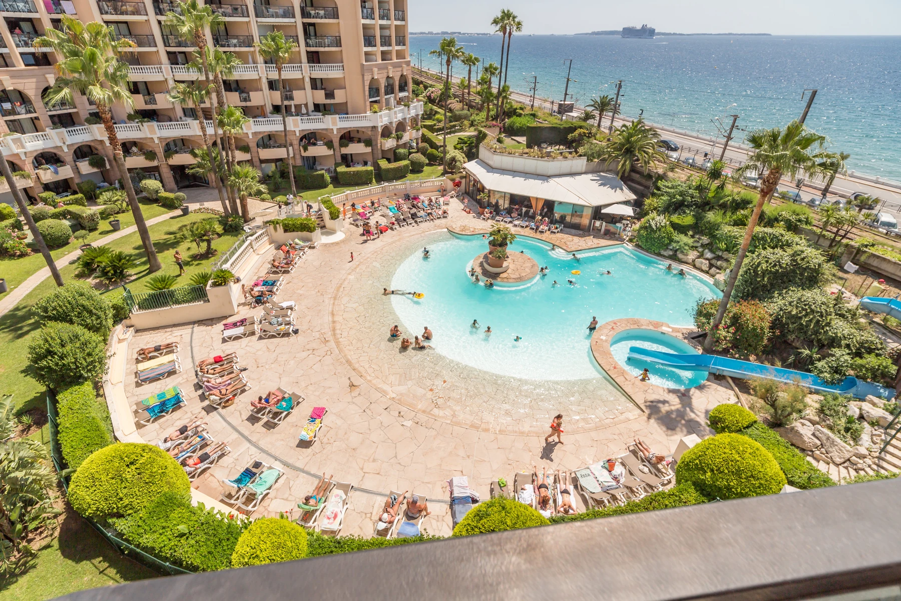 Guests lounge on poolside chairs and swim in a resort's pool; a seaside backdrop completes the sunny scene at Cannes Verrerie
