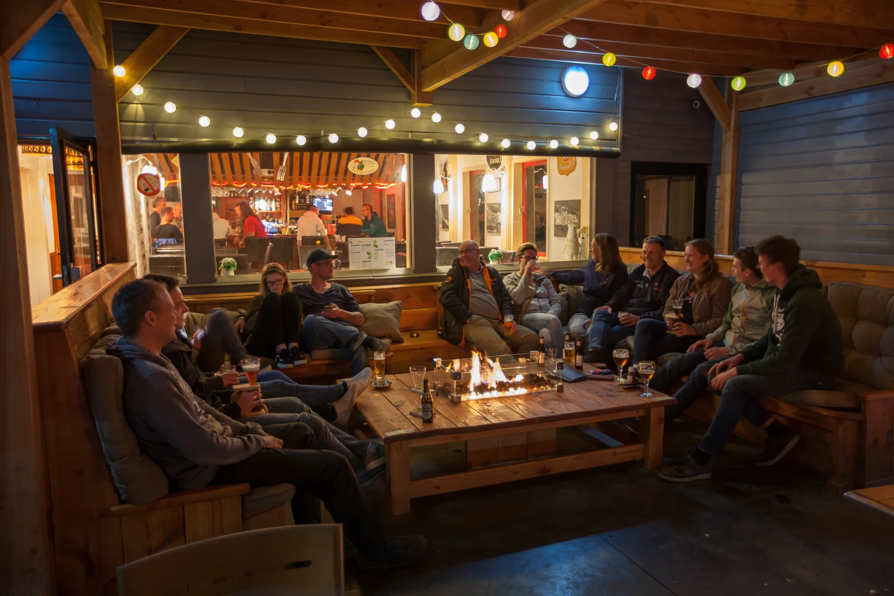 A group of people sitting around a fire pit, socializing under festive string lights in a cozy patio area at Polleur