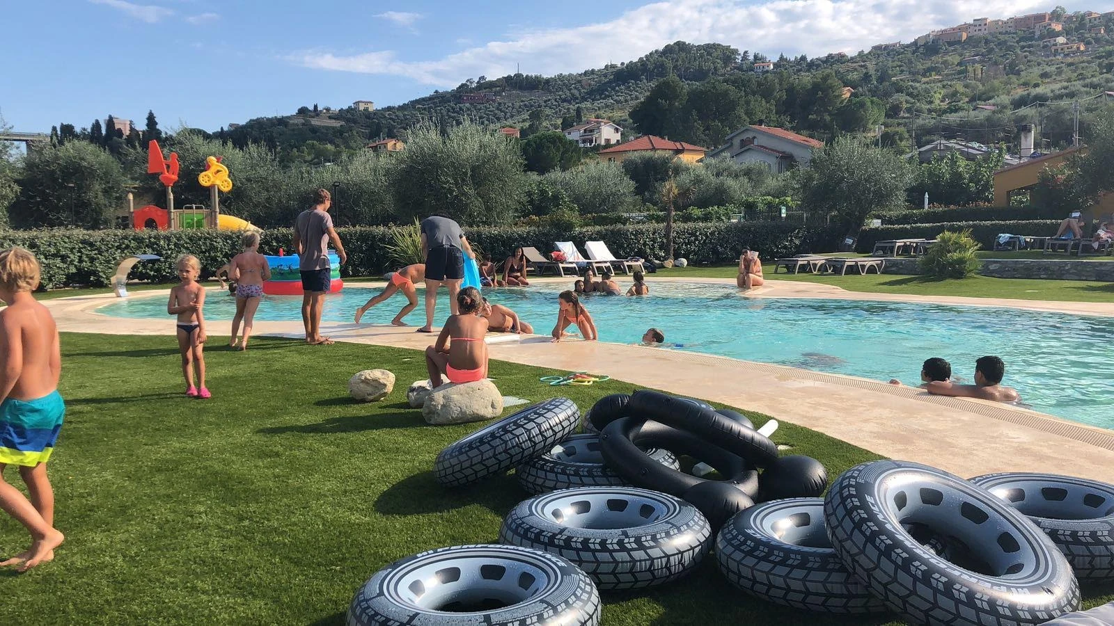 Children and adults playing and relaxing in and around a pool, surrounded by greenery and hills at Residence Rebi Village