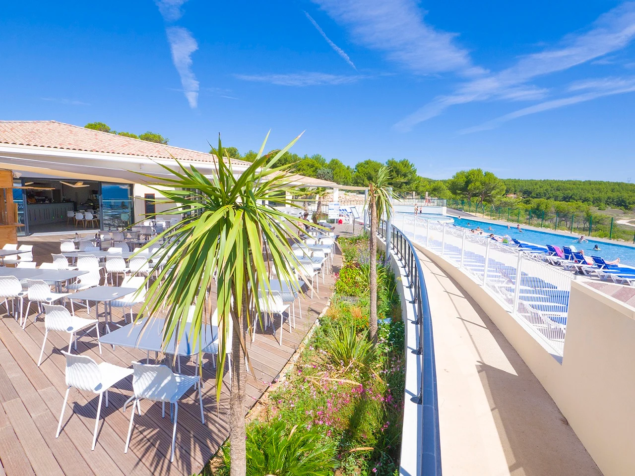 Tables and chairs are arranged on a wooden deck near a pool area, surrounded by trees and blue skies at L'Arquet - Côte Bleue