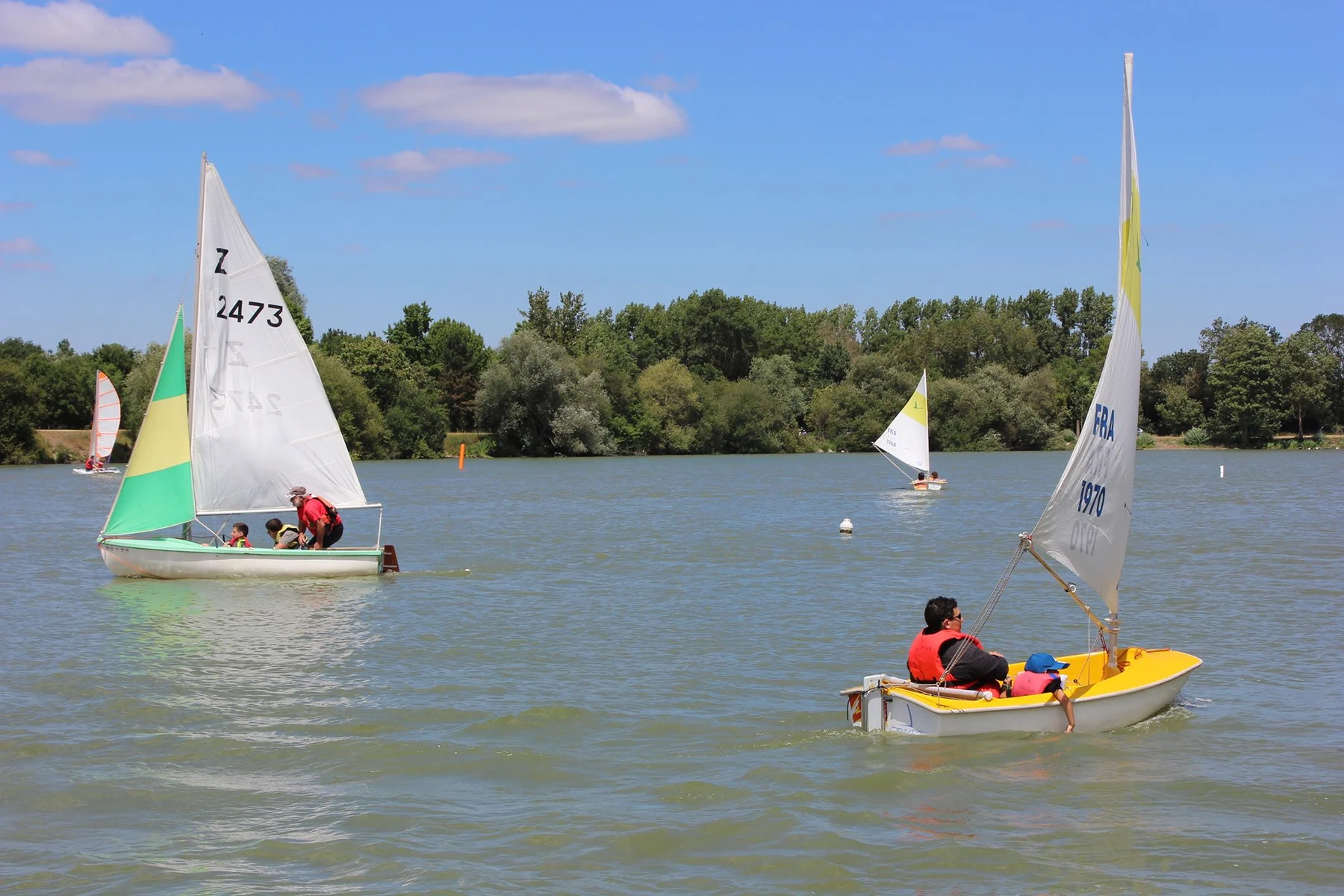 Sailboats navigating a lake surrounded by trees under a blue sky with scattered clouds at Seasonova Du Chêne