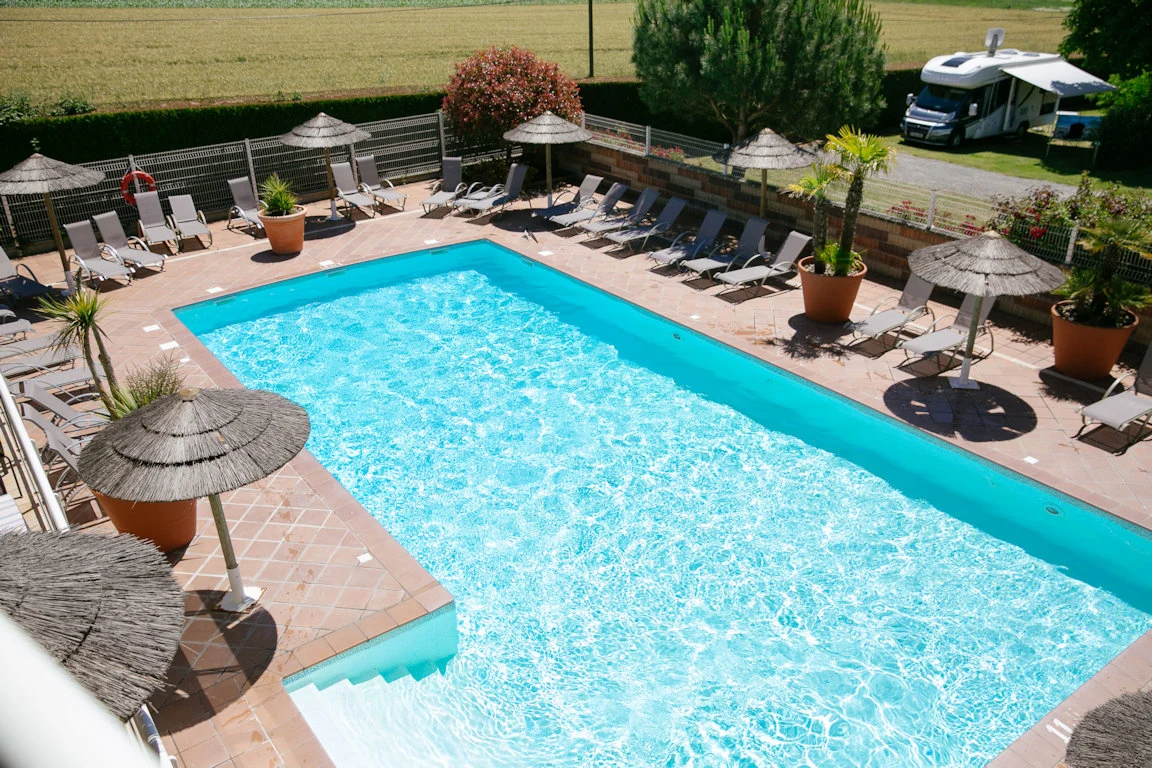 Swimming pool with clear water is surrounded by lounge chairs, parasols, and potted plants, near a parked RV at Seasonova Saint-Michel