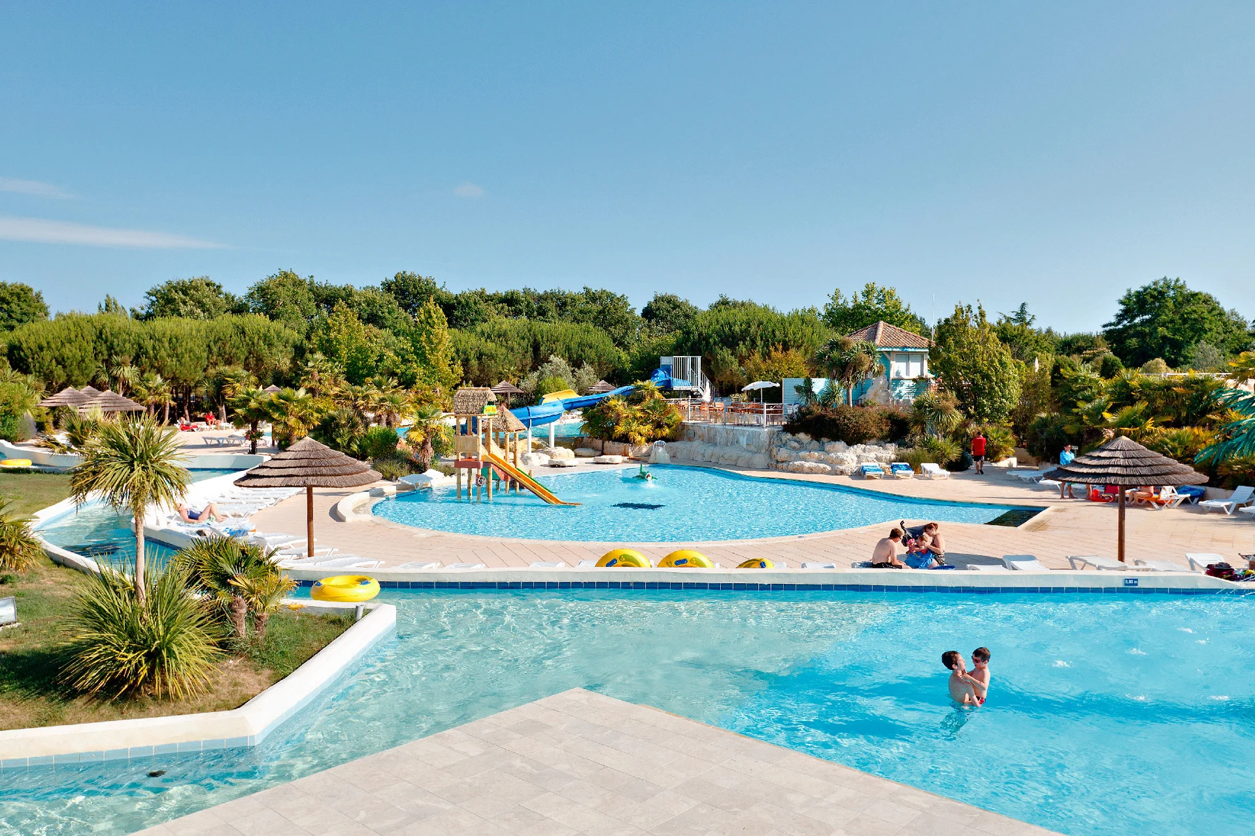 People swim and play in a large outdoor pool complex surrounded by greenery and shaded lounging areas at Sequoia Parc