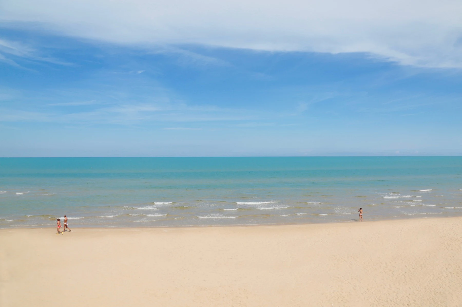 Two people walking on a sandy beach with gentle waves under a clear blue sky at San Menaio