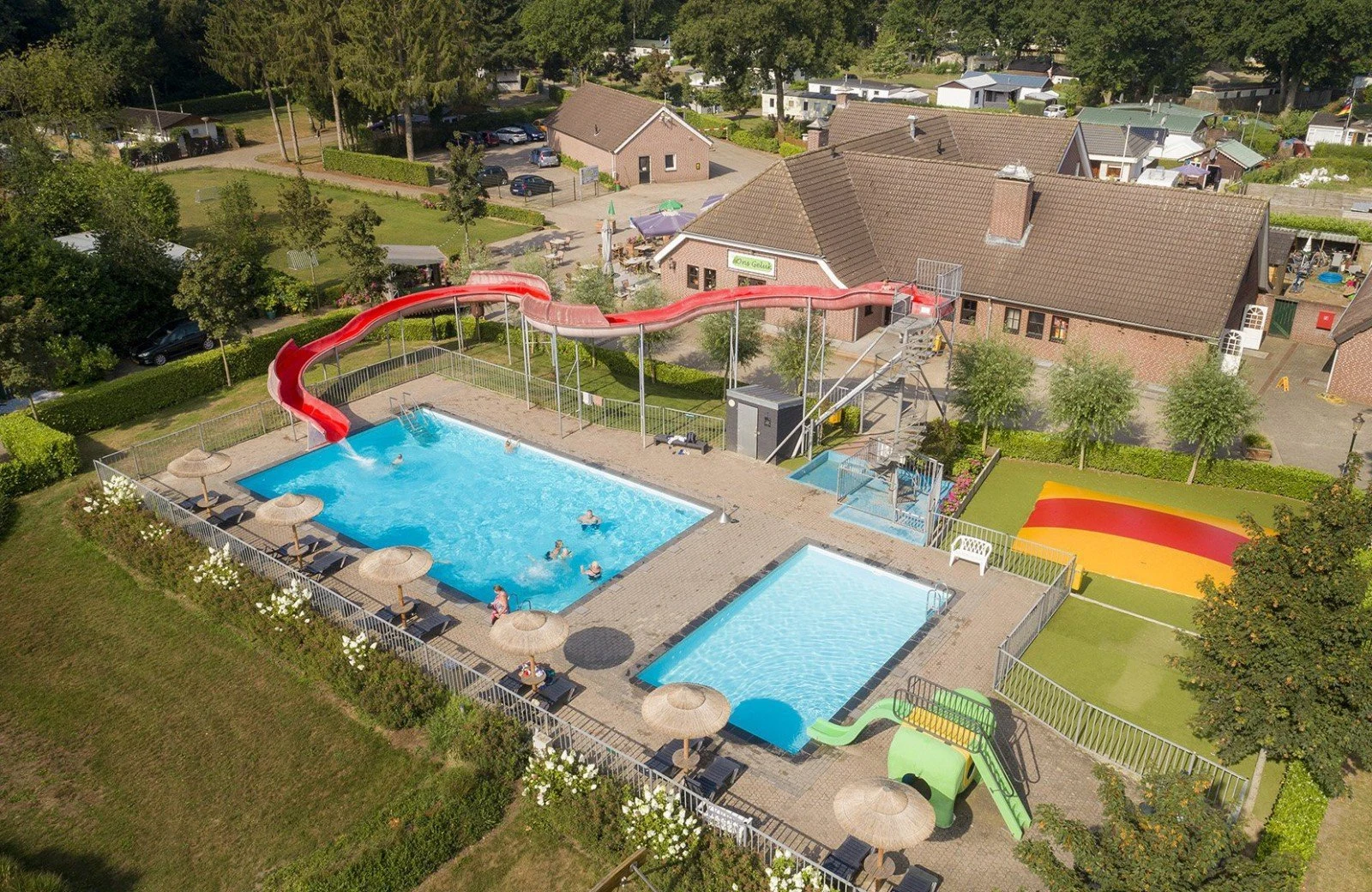 Swimming pool with waterslide, occupied by people, situated next to a large building and camping area surrounded by greenery at Hoeve de Schaaf
