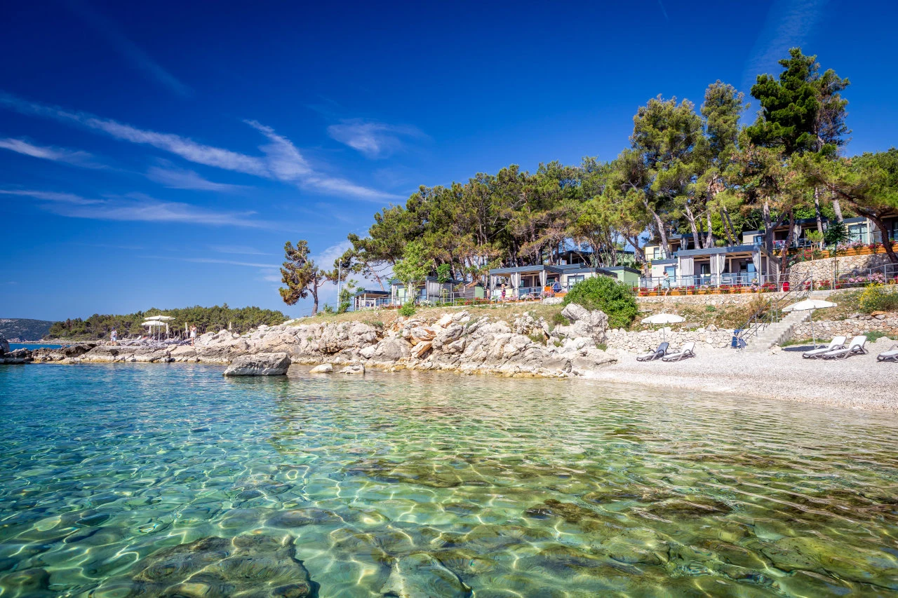 Clear turquoise water lapping against rocky shore below a hillside covered with pine trees and cabins at Ježevac Premium Camping Resort
