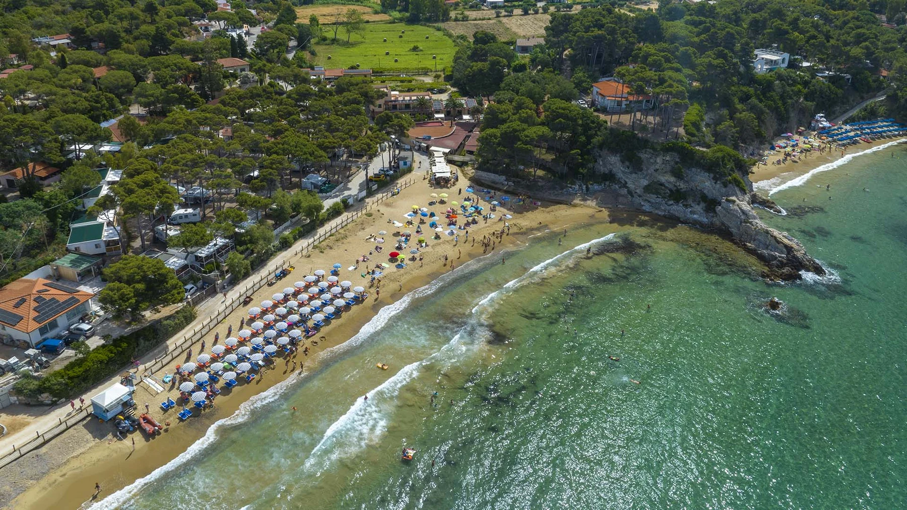 Beach umbrellas shade sunbathers on a sandy shore, waves gently crashing, surrounded by forested hills and beachfront structures at Europa Camping Village Elba