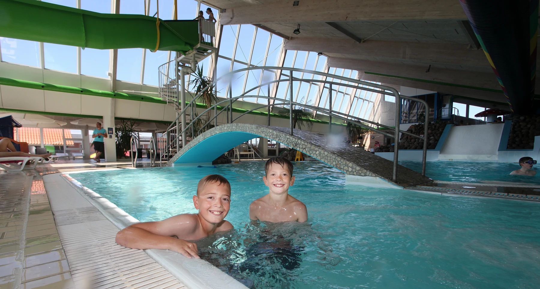 Two children smile while swimming in an indoor pool, underneath a small arched bridge at Dancamps Nordsø