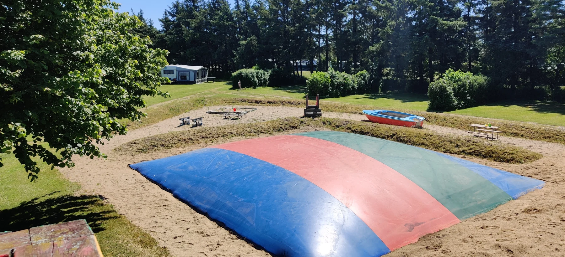 A colorful inflatable jumping pad in a sandy area surrounded by picnic tables, a small boat, and trees at Ballum