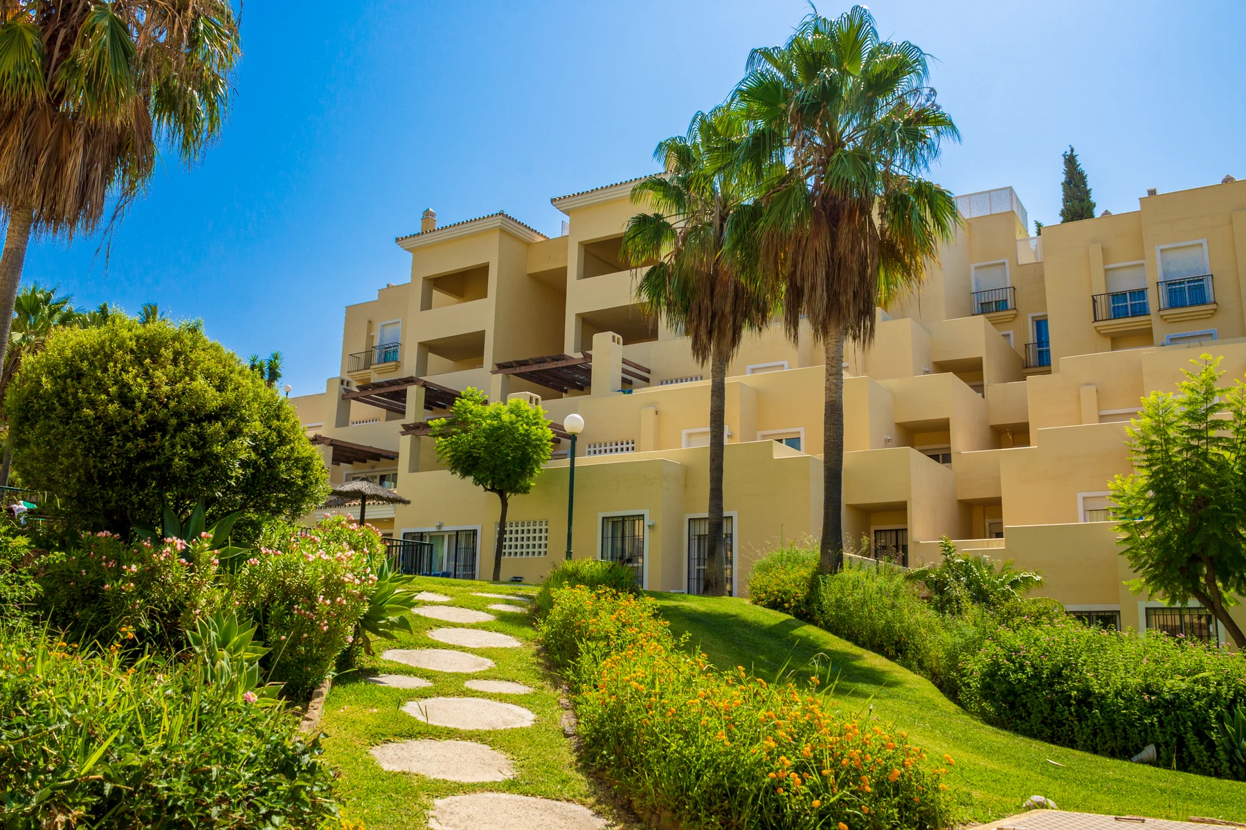Modern, beige apartment building surrounded by lush tropical landscaping and a stone path, under a clear blue sky at Colina del Paraiso
