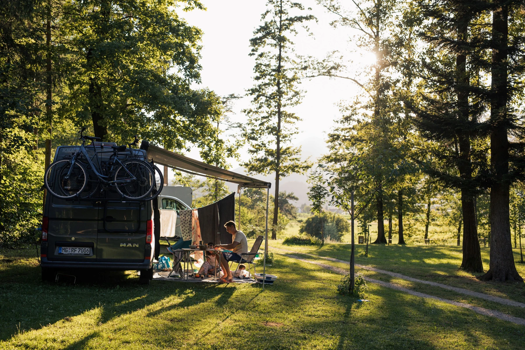 Campervan with bicycles on top, people sitting and relaxing under an awning, surrounded by lush trees at Forest camping Mozirje