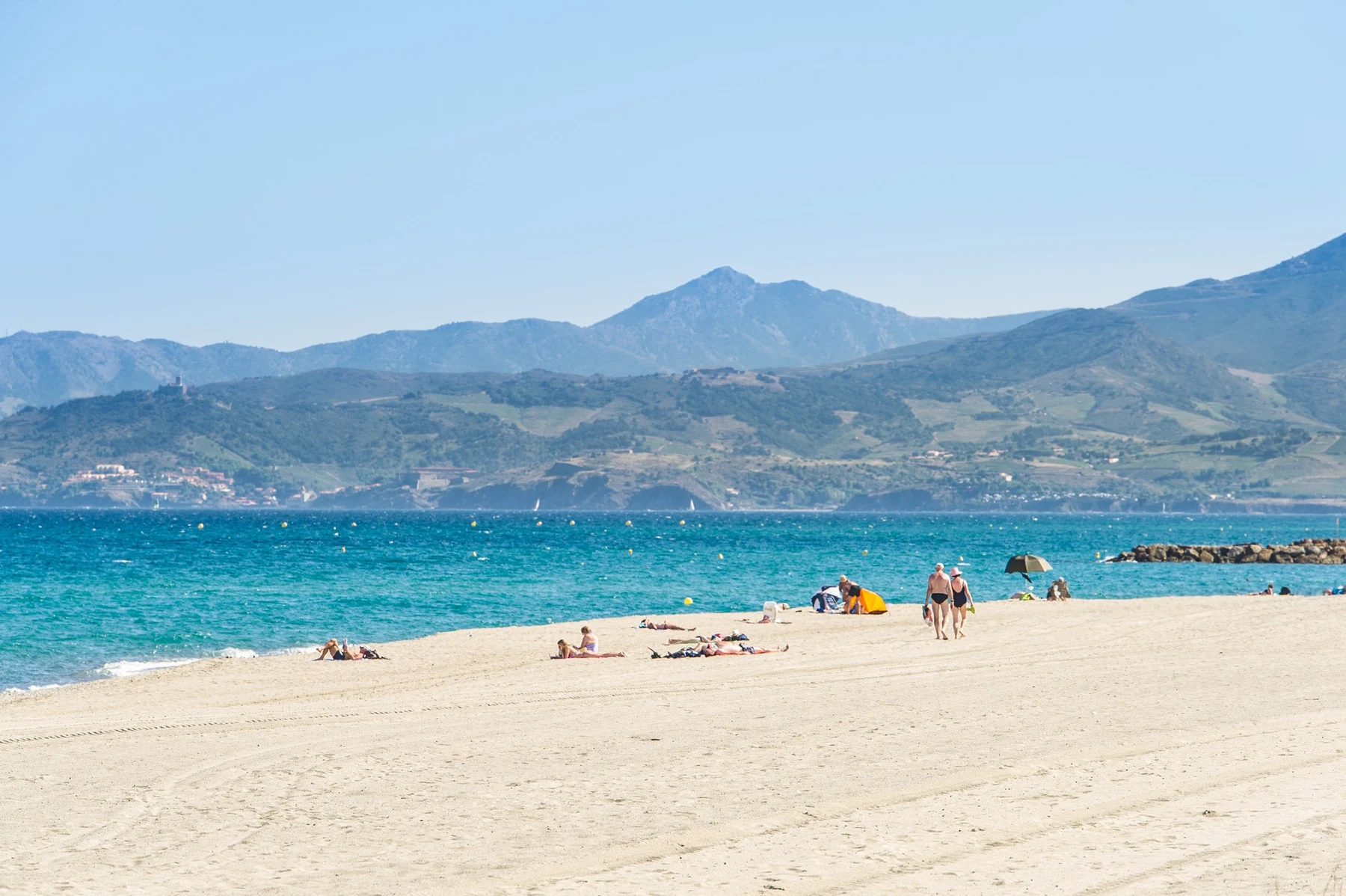 People relax on a sandy beach near turquoise water with distant hills and mountains at Marvilla Parks Le Soleil de la Mediterranee