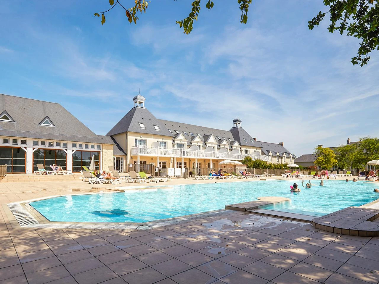 Swimming pool with people relaxing and swimming, surrounded by deck chairs and a large building with multiple windows at Le Green Beach