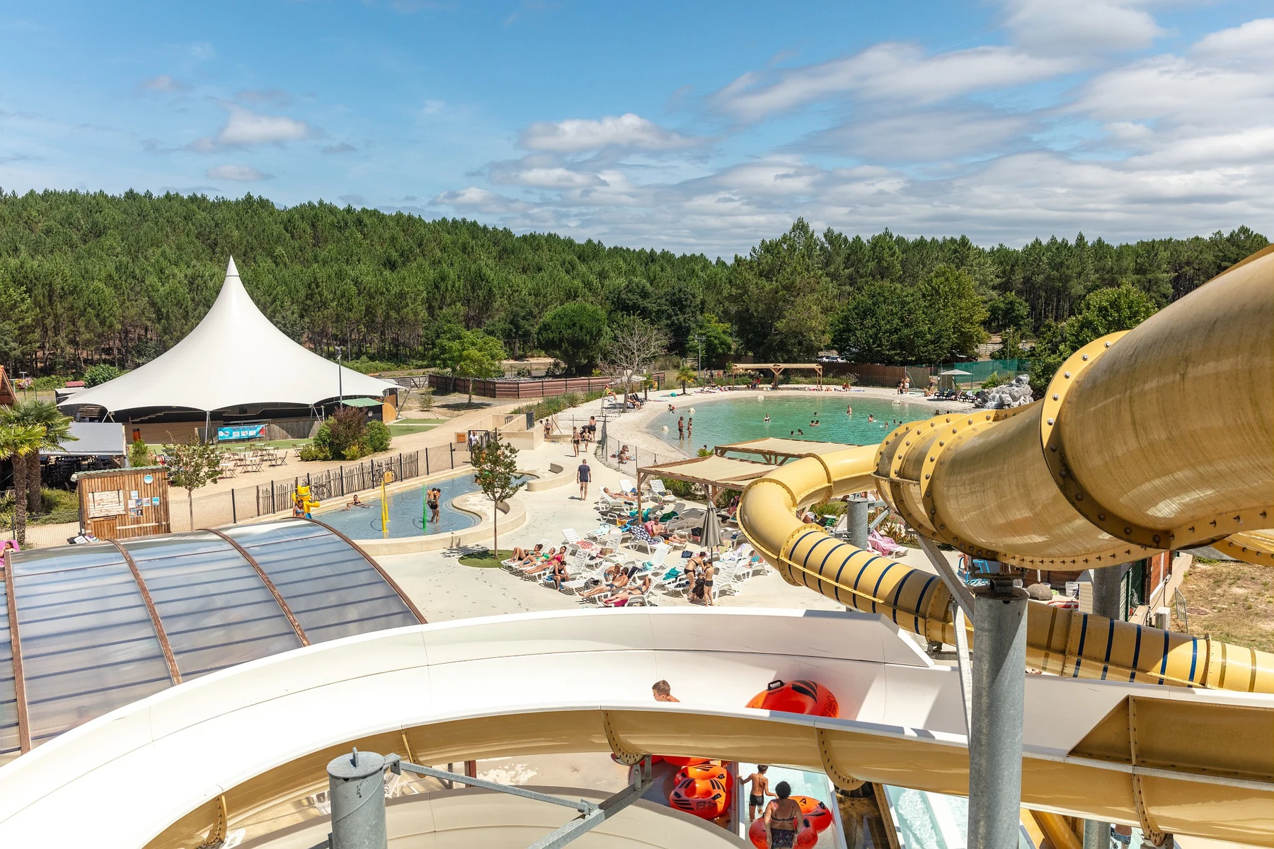 Curved waterslide descends towards a busy pool area with lounging people surrounded by trees at Marvilla Parks Les Vignes