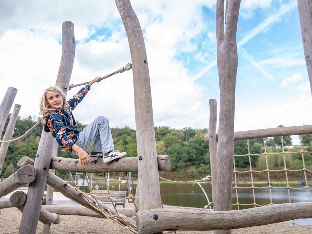 Child playing on a wooden jungle gym beside a lake, surrounded by trees and cloudy sky at Landal Glamping Neufchâteau