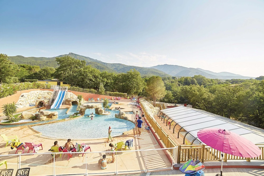 Swimming pool surrounded by people playing and relaxing; mountains and trees in the background at Les Albères