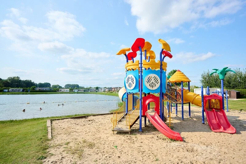 Colorful playground structure with slides and ladders on sandy ground, adjacent to a lake with swimmers at Betuwe